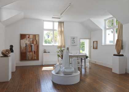 The first floor of the museum. The room is bright, with a large window and open door leading to the garden. There are large white sculptures on the wooden floor and paintings hanging on the walls.