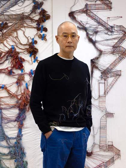 A portrait of artist Do Ho Suh in his studio. He is surrounded by sculptures made with blue and red threads.
