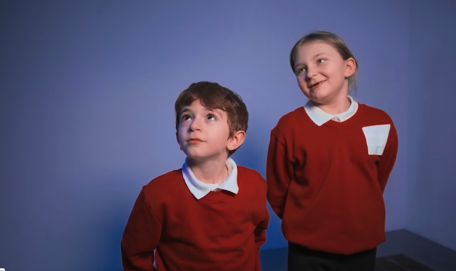 Two children in red jumpers look up at something off camera, standing against a blue wall